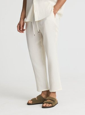 Persona che indossa pantaloni testurizzati color ecrù e camicia abbinata con sandali a doppio cinturino verde oliva su sfondo chiaro.