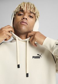 Beige színű kapucnis pulóver zsinórokkal és fekete részletekkel, az elején egy fekete Puma logóval. Puha anyag és laza fit.