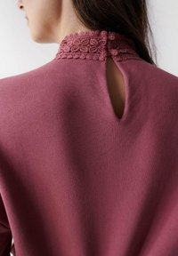 Top color rosa con un alto cuello de encaje que presenta un patrón floral, detalle de ojal en la espalda y cierre de botón en el cuello. Textura de tela suave.