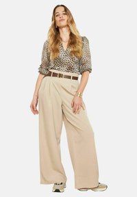 Bēša plaša stila bikses ar krokojumu, kombinētas ar leoparda raksta blūzi un brūnu jostu. Modelis valkā sporta apavus un ir ar gariem viļainiem matiem.