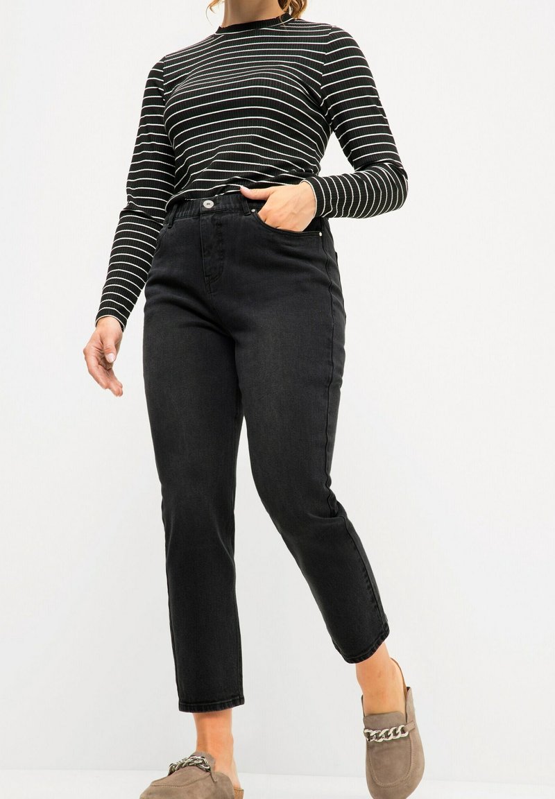 Femme portant un jean noir taille haute, un haut à manches longues rayé noir et blanc, et des chaussures beige à enfiler avec un détail en chaîne métallique.