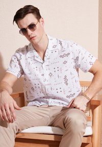 Korte mouwen witte shirt met geometrische bruine patronen, gecombineerd met beige broek. Model zit op een houten stoel en draagt ronde zonnebril.