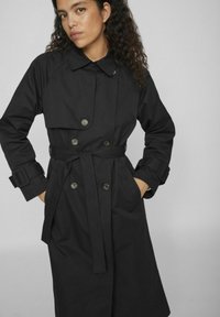 Cămașă neagră lungă cu un design cu două rânduri de nasturi, guler mare, talie cu curea și buzunare laterale. Fabricată dintr-un material neted, rezistent la apă.