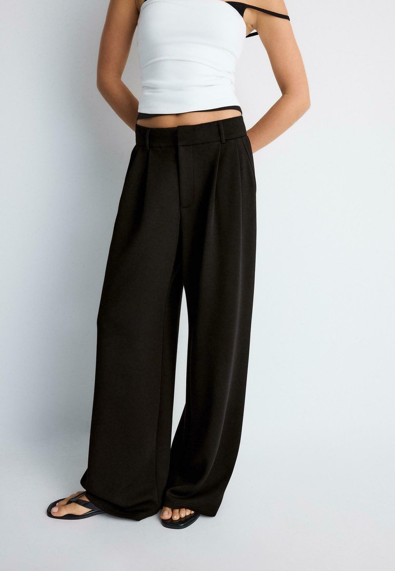 Femme portant un pantalon large noir ample, un crop top blanc sans manches avec bretelle noire et des tongs noires sur fond uni.