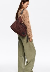 Brązowa pleciona torebka o teksturowanym, siateczkowym wzorze i kontrastującym brązowym wnętrzu. Posiada pasek na ramię oraz małą naszywkę z logo.