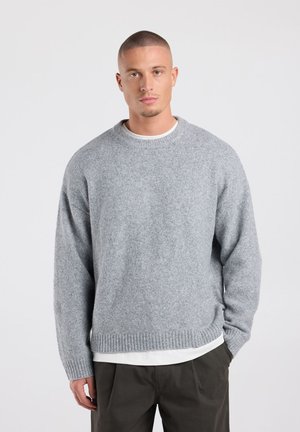 Jonge man met kort haar die een grijze gebreide trui draagt over een wit overhemd en een donker broek, staand tegen een eenvoudige lichte achtergrond.