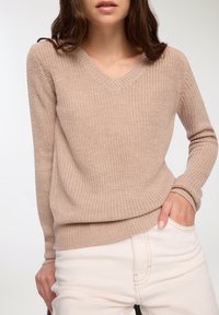 Beżowy dzianinowy sweter z dekoltem w kształcie V, długimi rękawami i prążkowanymi mankietami. Noszony na jasnych spodniach, prezentujący teksturowany, casualowy design.