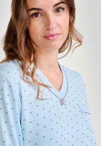 Joven mujer con ojos marrones y cabello ondulado lleva una blusa azul claro con lunares y un pequeño lazo cerca del cuello en V.