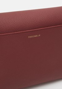 Μπορντό δερμάτινο πορτοφόλι με υφή, που διαθέτει το χρυσό ανάγλυφο λογότυπο "COCCINELLE" στη μια πλευρά και λείο φινίρισμα στην επάνω επιφάνεια.