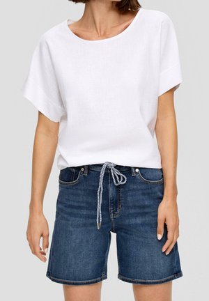 Kvinde iført en plain hvid kortærmet top, stoppet ned i mørkeblå denimshorts med et sort-hvidt reb bælte bundet i taljen.