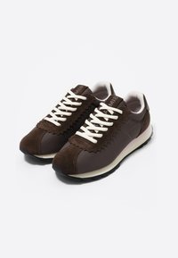 Un par de zapatillas marrones con cordones blancos, un cuello acolchado suave, acentos de ante y suelas negras texturizadas sobre fondo blanco.