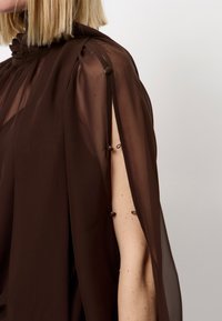 Bruine sheer stof top met hoge hals, gestructureerde schouders en open mouwen, vastgemaakt met decoratieve knopen langs de arm.