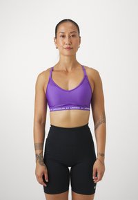 Sujeción deportiva morado con tirantes finos, que presenta una banda blanca ancha con la inscripción "Under Armour". Combinado con pantalones cortos negros de tiro alto.