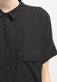 La camicia nera a maniche corte presenta un colletto a punta, bottoni frontali e una tasca applicata, realizzata in un tessuto testurizzato.