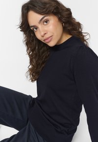 Jūras spēku zila adīta džemperis ar augstu kakla līniju, pūkainām piedurknēm un ribinātu apakšmalu; mīksta tekstūra un pieguloša forma. Apvienots ar tumšiem biksēm.