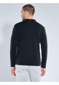 Schwarzes Langarmoberteil aus strukturiertem Stoff mit Rundhalsausschnitt, seitlichen Schlitzen und kleinem Logo am Nacken hinten.