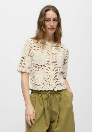 Kvinne iført en kortermet, kremfarget heklet cardigan med blomster mønster og olivengrønne bukser med snøring, stående med den ene hånden i lommen.