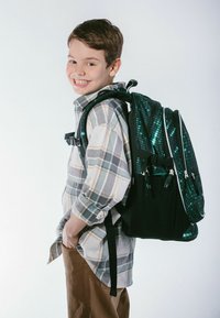 Zaino nero con accenti olografici verdi e motivi digitali, portato da un bambino in una camicia a quadri e pantaloni marroni.