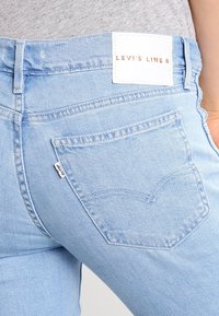 Jeans de mezclilla azul claro con una vista en primer plano del bolsillo trasero, que presenta detalles de costura y una etiqueta blanca de marca en la cintura.
