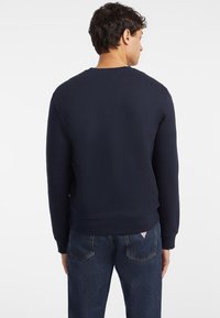 Tengerészkék crewneck pulóver bordázott textúrával és hosszú ujjakkal, kék farmerral, hátsó zsebében címkével.