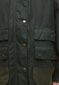 Veste vert foncé à l'avant avec quatre poches à rabat, boutons-pression en métal et nom de marque Barbour visible sur la poche inférieure droite.