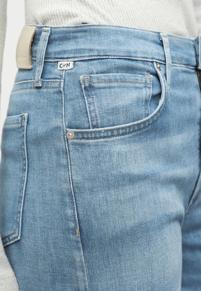 Κοντινή λήψη από ανοιχτό μπλε τζιν με μπροστινή τσέπη, θηλιά ζώνης, κουμπί και μικρή λευκή ετικέτα κοντά στη μέση που φοριέται με ανοιχτό γκρι ριμπ πλεκτό τοπ.