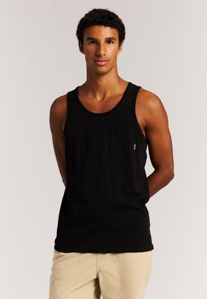 Junger Mann mit kurzen lockigen Haaren, der ein schwarzes Tanktop mit Brusttasche und beige Hose trägt, steht vor einem schlichten hellen Hintergrund.