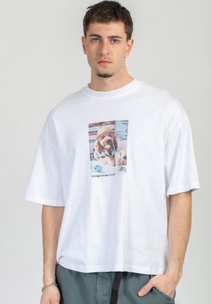 Giovane uomo che indossa una maglietta bianca oversize con una foto stampata di un cane con cappello che sorseggia una bevanda in un bar.