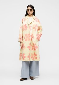 Γυναίκα με γυαλιά ηλίου, μακρύ μπεζ παλτό με μεγάλα ροζ λουλουδένια σχέδια, φαρδιά ανοιχτό μπλε παντελόνια και καφέ παπούτσια, στέκεται μπροστά σε λευκό φόντο.