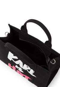 Sac fourre-tout noir en toile avec une poche zippée, des compartiments intérieurs et un design de texte audacieux en blanc et rose d'un côté. Deux poignées robustes.