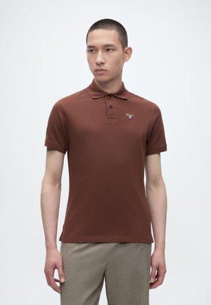 Junger Mann steht, trägt ein braunes kurzärmeliges Poloshirt mit kleinem Logo auf der Brust und hellgraue Hose.