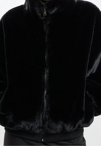 Černá bunda z umělé kožešiny s předním zapínáním na zip, vysokým límcem a žebrovanými manžetami. Měkká textury a plyšový vzhled. Zaoblený lem.