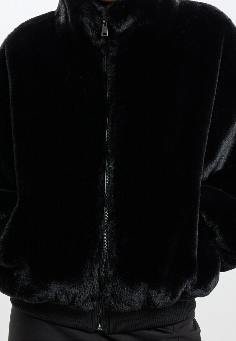 Černá bunda z umělé kožešiny s předním zapínáním na zip, vysokým límcem a žebrovanými manžetami. Měkká textury a plyšový vzhled. Zaoblený lem.