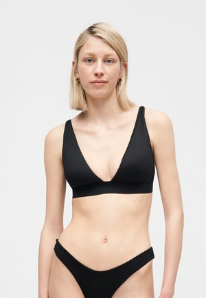 Femme blonde portant des boucles d'oreilles créoles dorées et un bikini noir avec un haut en V profond et un bas taille haute, se tenant devant un fond clair uni.
