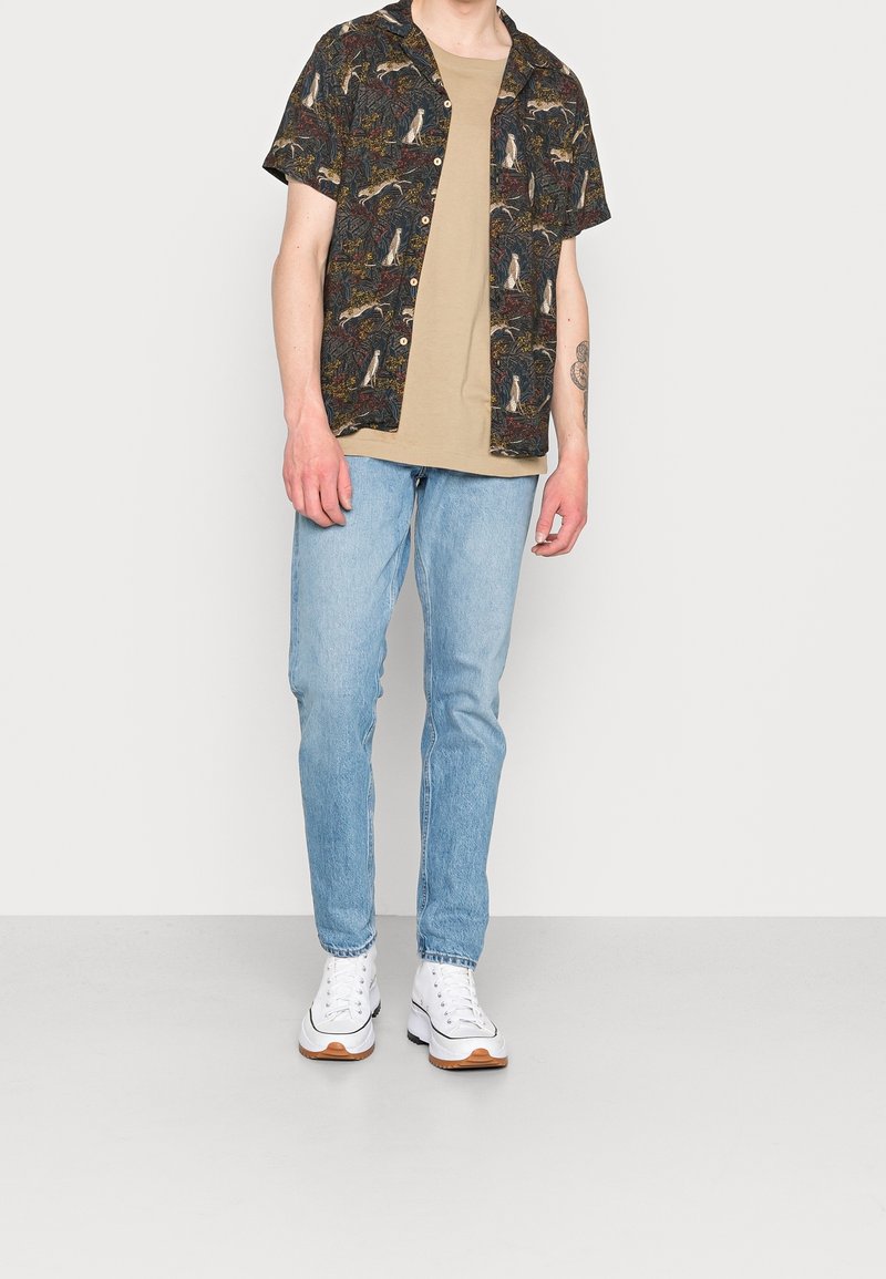 Camicia a maniche corte a motivo scuro con bottoni, abbinata a una maglietta beige, jeans azzurri chiari e sneaker bianche con dettagli marroni.