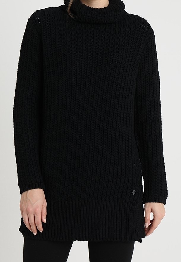 Femme portant un pull col roulé noir en maille côtelée, long avec manches longues et fentes latérales, associé à un pantalon noir.