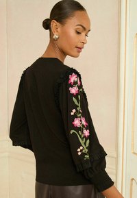 Fekete felső rózsaszín és zöld virágmotívummal az egyik ujjon, fodros részletekkel és puffos ujjvégekkel. Simára anyag textúra.