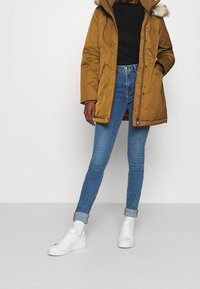 Bruine parka-jas met een met bont gevoerde capuchon, ritssluiting, twee zijzakken, gedragen over een zwarte top en blauwe jeans, gecombineerd met witte sneakers.