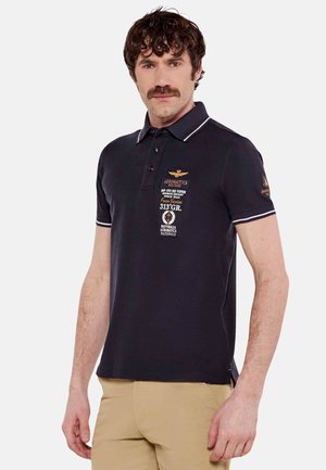 Polo shirt nautico in cotone con accenti bianchi. Presenta loghi e testi ricamati sulla parte anteriore. Maniche corte e design classico del colletto.