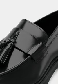 Mocassins en cuir noir à finition brillante, avec un nœud décoratif à l'avant, une texture lisse et des accents de coutures contrastantes.