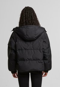 Persona con capelli lunghi intrecciati che indossa un giubbotto imbottito nero con cappuccio, in piedi di spalle rispetto alla fotocamera su uno sfondo semplice.