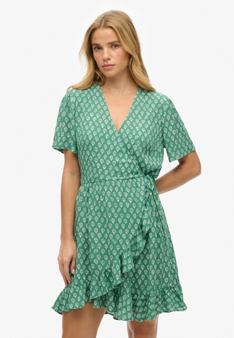 Vestido verde envolvente con mangas cortas, que presenta un patrón floral, un dobladillo con volantes y un detalle de lazo en la cintura. Tela ligera y texturizada.