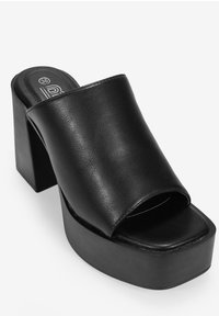 Mule negro con tacón de plataforma, que presenta una amplia banda frontal y un tacón cuadrado grueso. Textura de cuero suave con un diseño minimalista.