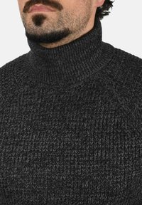 Jersey de cuello alto gris oscuro con un patrón de tejido texturizado. Presenta un cuello alto y un diseño de mangas raglán, ofreciendo una silueta ajustada.