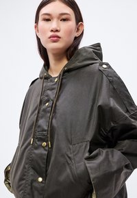 Must suurusega parka kapuutsiga, millel on kuldsed metalldetailid, tõmblukuga sulgur ja suured taskud. Sile tekstuur, mati viimistlusega.