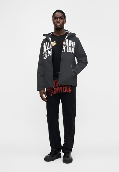 Μαύρο μπουφάν με κουκούλα και λευκό κείμενο, φορεμένο πάνω από μαύρο t-shirt και κόκκινο παντελόνι με γραφικό σχέδιο. Μαύρα αθλητικά παπούτσια ολοκληρώνουν την εμφάνιση.