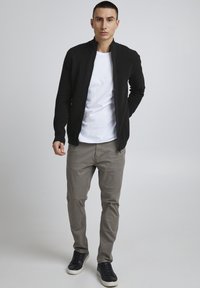 Schwarz gerippten Reißverschlussjacke über einem weißen T-Shirt, kombiniert mit hellgrauen Hosen und schwarzen Sneakers. Einfaches und modernes urbanes Outfit.