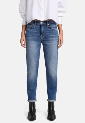 Jean skinny en denim bleu moyen, avec une finition délavée, des ourlets roulés et un style à cinq poches, associé à une chemise blanche à manches longues.