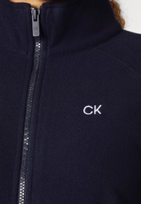 Närbild av en marinblå fleecejacka med frontdragkedja och vitt broderat "CK"-logotyp på bröstet, bärs av en person.