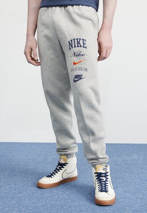 Osoba nosząca jasnoszare spodnie dresowe Nike z wieloma logo Nike na jednej nodze oraz białe buty sportowe Nike z wysoką cholewką i ciemnymi sznurowadłami.